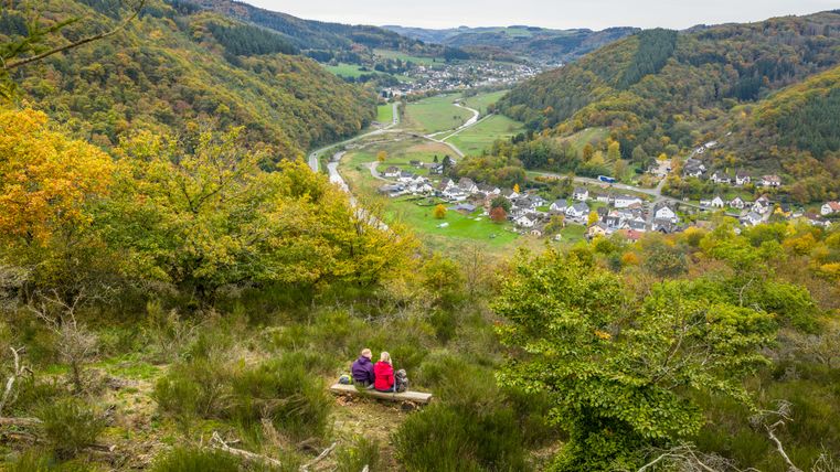Zwei Personen sitzen auf einer Bank am Hang und blicken auf ein herbstliches Tal mit einem kleinen Dorf und einem Fluss