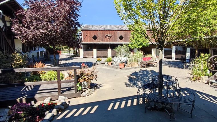 Une cour pittoresque avec des plantes et des arbres en fleurs. Au centre se trouve un coin salon et en arrière-plan, des bâtiments historiques sont visibles.