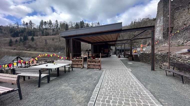 Een modern restaurantgebouw met een geplaveide patio temidden van een natuurlijke omgeving. Op de achtergrond zijn ruïnes en gekleurde vlaggen te zien.
