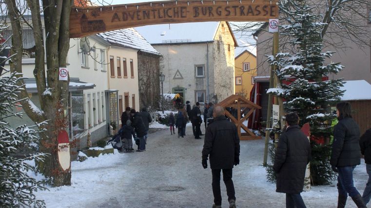 Eine verschneite Straße mit einem Holzbogen an der Einfahrt und Menschen, die spazieren gehen. Die Häuser sind im winterlichen Stil dekoriert.