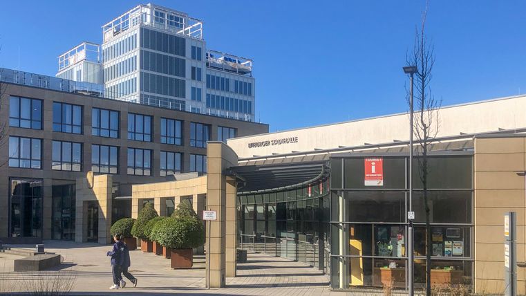 Ein modernes Gebäude mit großen Fenstern und einer klaren Architektur. Im Vordergrund ist eine Person zu sehen, die einen Gehweg entlanggeht.