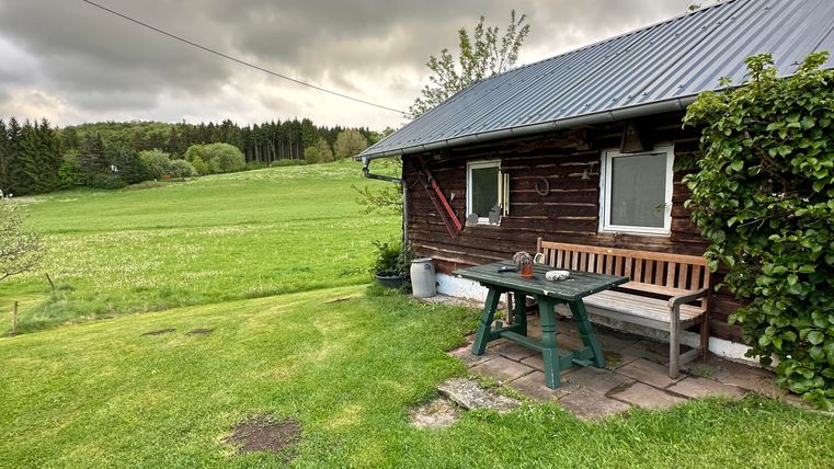 Ein gemütliches Holzhaus steht in einer grünen Wiese. Der Himmel ist bewölkt, und es gibt einen Tisch mit einer Bank vor dem Haus.