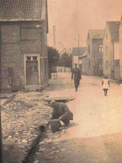 A historic street scene with cobblestones and old houses. A man is working on the ground while a person and a child walk by.