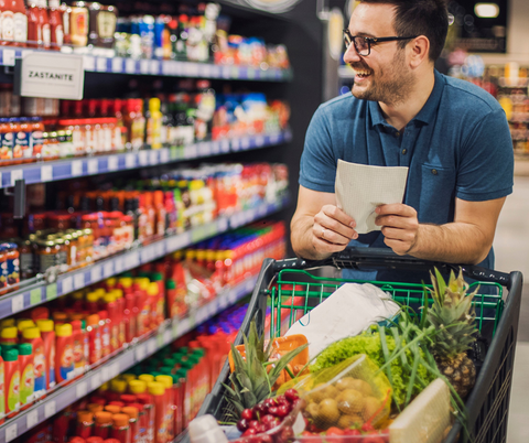 <p>Een man met een goed gevulde winkelwagen loopt door een gang in de supermarkt. In de schappen zijn sauzen en ketchup te zien. In zijn hand houdt hij een boodschappenlijst.</p>