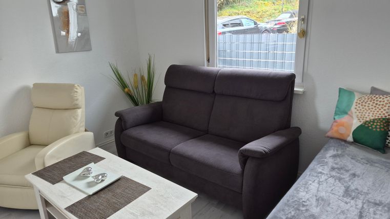 Een gezellige woonkamer met een donkergrijze bank en een lichte fauteuil. De tafel is voorzien van een tafellaken en decoraties, en het raam biedt uitzicht naar buiten.