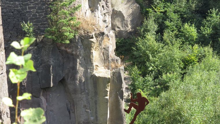 A climber hanging from a steep rock face, surrounded by green vegetation. Rocky formations are visible in the background.