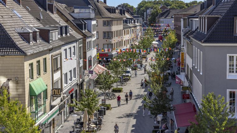 Een drukke winkelstraat met bomen en winkels. Veel mensen slenteren langs de straat.