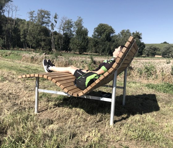 Eine Person entspannt auf einer geschwungenen Holzbank in einer grünen Landschaft unter blauem Himmel., © Tourist Information Bitburger Land