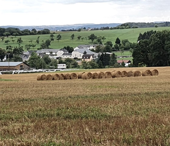 View of a field with bales of straw, a village and green hills in the background. The sky is cloudy., © TI Bitburger Land - Steffi Wagner