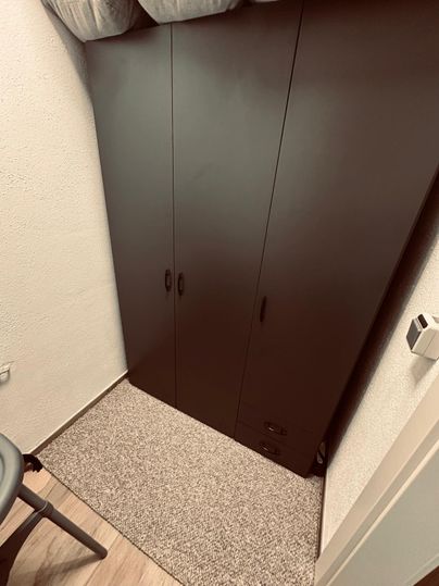 Ein schmaler, schwarzer Kleiderschrank steht an der Wand. Der Boden ist mit einem hellen Teppich ausgelegt.