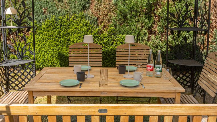 Een mooie houten tuintafel met stoelen en gedekte borden. Op de achtergrond zijn groene planten te zien.