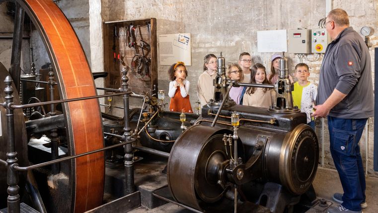 Eine Gruppe von Kindern hört einem Erklärer zu, der eine alte Maschine zeigt. Die Maschine besteht aus einem großen Schwungrad und einem metallischen Antrieb.