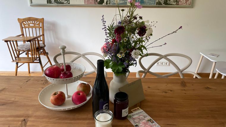 Une belle table dressée avec un vase plein de fleurs et divers aliments. Sur la table, il y a des pommes, une bouteille de vin et un bougeoir.