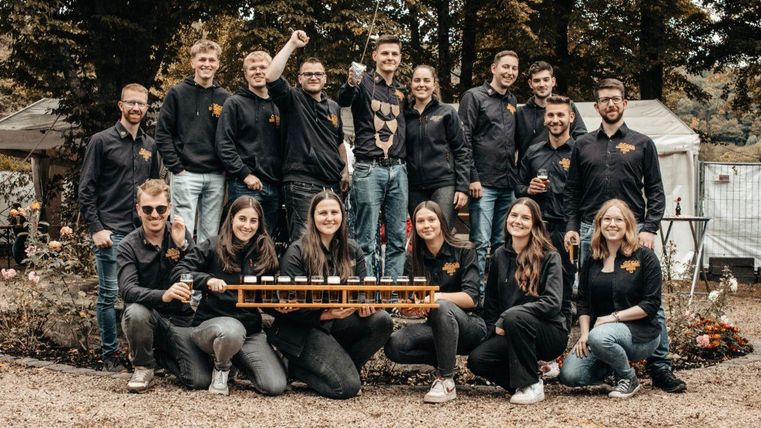 Eine Gruppe von jungen Menschen steht lächelnd auf einer Wiese. Sie machen Daumen hoch und präsentieren eine Partie Getränke auf einem Tablett.