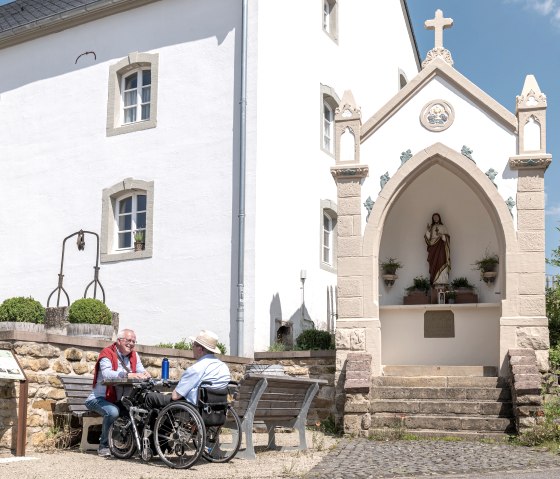 Zwei Männer, einer im Rollstuhl, sitzen auf einer Bank vor einem weißen Gebäude mit einer religiösen Statue in einer Nische. Sonniger Tag., © Naturpark Südeifel, Thomas Urbany