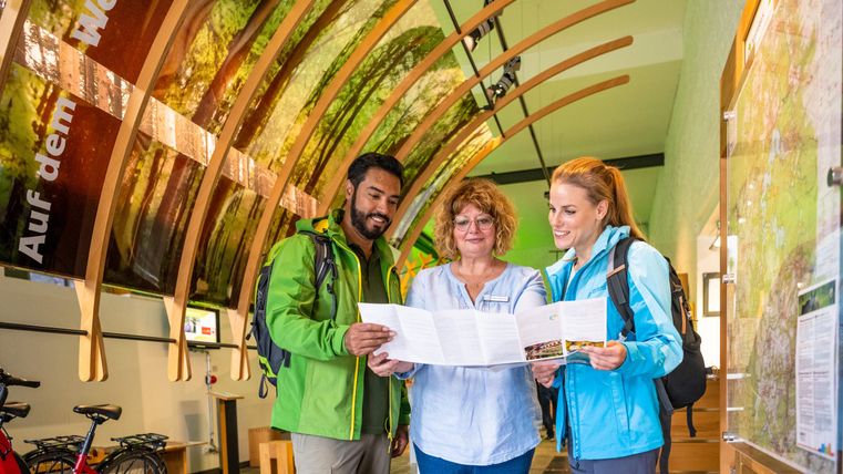 Three people are standing in an information center and looking at a map. In the background, bicycles and a wooden ceiling with plant patterns can be seen.