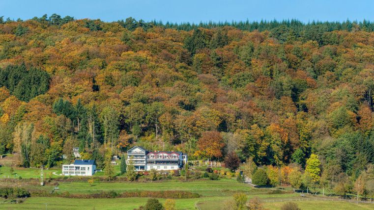 Een pittoreske vallei met kleurrijke bomen in de herfst. Op de voorgrond staan twee gebouwen, omgeven door de natuur.