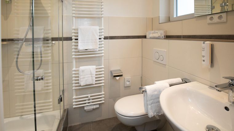 A modern bathroom with a shower, sink, and towel rack. The walls are light and the fixtures are clean and elegant.