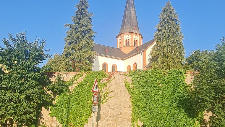 Eine kleine Kirche auf einem Hügel, umgeben von Bäumen und Sträuchern. Der Himmel ist klar und blau.