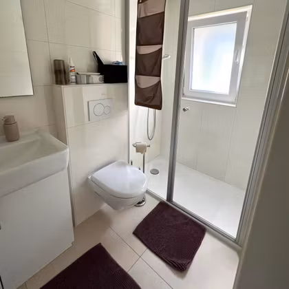 Une salle de bains moderne avec douche, toilettes et lavabo. Deux tapis de bain violets sont placés sur le sol.