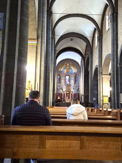 Eine Innenaufnahme einer Kirche mit hohen Decken und Holzbankreihen. Zwei Personen sitzen auf Bänken und blicken nach vorne.