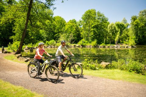 Ein Radfahrer-Paar sitzt auf einem Baumstumpf und entspannt sich. Im Hintergrund gibt es einen Radweg und viel grüne Landschaft.