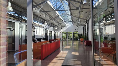 Ein moderner, heller Innenraum mit großen Fenstern und einem Glasdach. Verschiedene Sitzbereiche und Pflanzen schaffen eine einladende Atmosphäre.