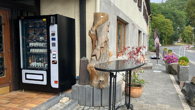 Ein Verkaufsautomat steht vor einem Gebäude, daneben befindet sich ein Tisch aus Metall und eine kunstvolle Holzskulptur. Im Hintergrund sind Pflanzen und Bäume zu sehen, die eine gemütliche Atmosphäre schaffen.
