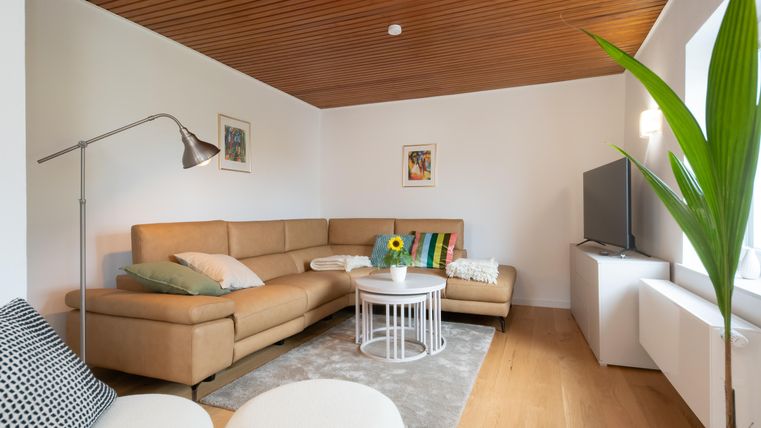 Ein modernes Wohnzimmer mit einem beigen Sofa und einem niedrigen Tisch. Die Wände sind hell gestrichen, und eine Pflanze steht in der Ecke.
