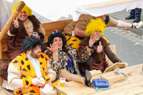 Eine Gruppe von Menschen in prähistorischen Kostümen sitzt fröhlich auf einem Holzfahrzeug. Sie lächeln und winken in die Kamera.