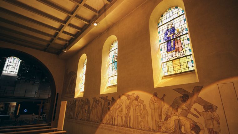 Der Innenraum der Kirche zeigt drei farbenprächtige Kirchenfenster. Zudem sind Gemälde an der Wand zu erkennen.