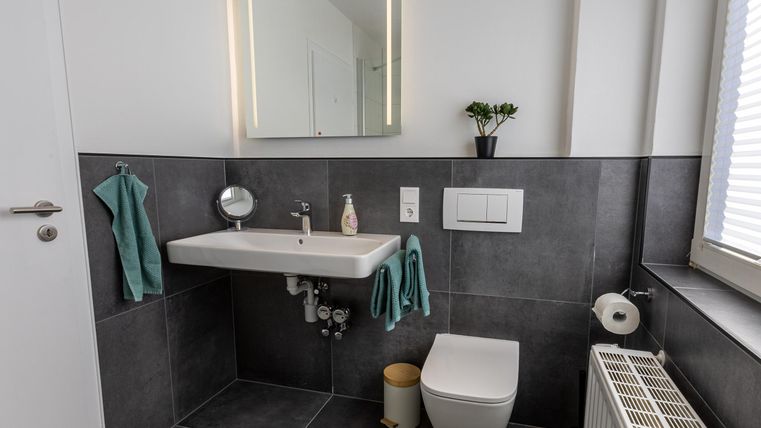 Een modern badkamerover een wandspiegel, een wastafel en een wandtoilet. De muren zijn donker betegeld en er zijn decoratieve handdoeken en planten.