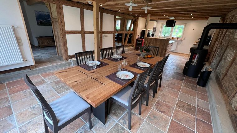 A cozy dining area with a large wooden table and chairs. The room has wooden beams and a modern kitchen in the background.