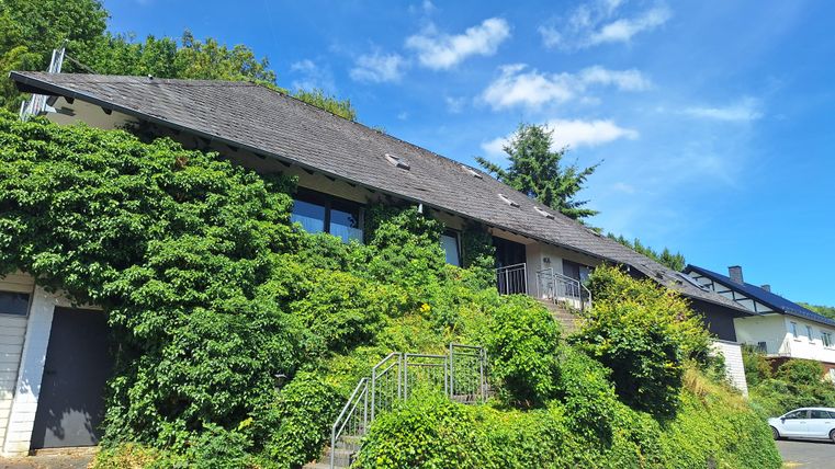 Une maison confortable, entourée de plantes vertes et d'arbres. Le ciel est bleu avec quelques nuages.