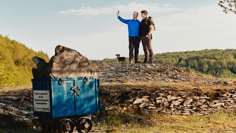 Twee wandelaars met een hond nemen een selfie op een leisteenveld in de Eifel. Links op de voorgrond staat een blauwe transportwagen met leisteen, daarachter bossen en groene heuvels onder een blauwe lucht.