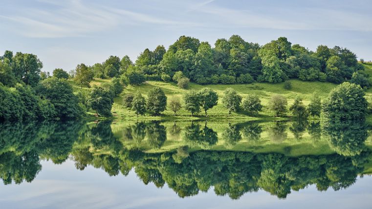 A quiet lake with mirrored water and lush greenery surrounding it. The trees and hills reflect beautifully in the water.