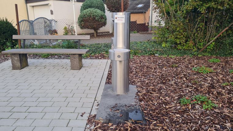 Een roestvrijstalen waterfontein staat in een klein park. In de buurt bevinden zich een bank en verzorgde bloembedden.