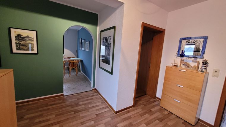 Ein einladender Flur mit einem grünen Akzentwand und Parkettboden. Auf der rechten Seite steht eine helle Kommode, während sich im Hintergrund ein Essbereich sichtbar ist.