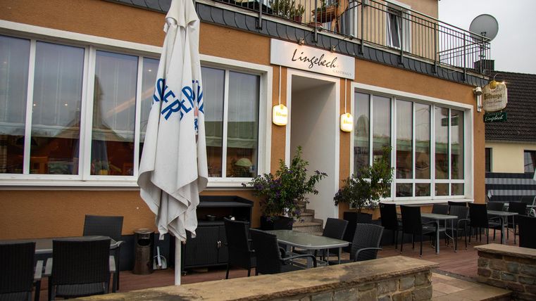 Een sfeervol restaurant met een terras en meerdere tafels. De façade is helder verlicht, en een parasol staat voor de ingang.