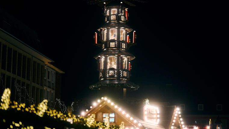 Ein festlich beleuchteter Weihnachtsbaum mit vielen Lichtern. Im Hintergrund sind gemütliche Holzstände zu sehen, die die winterliche Atmosphäre unterstreichen.