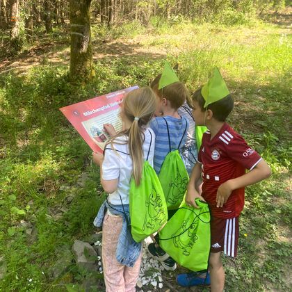 Drei Kinder mit grünen Rucksäcken und Hüten lesen ein Schild im Wald.
