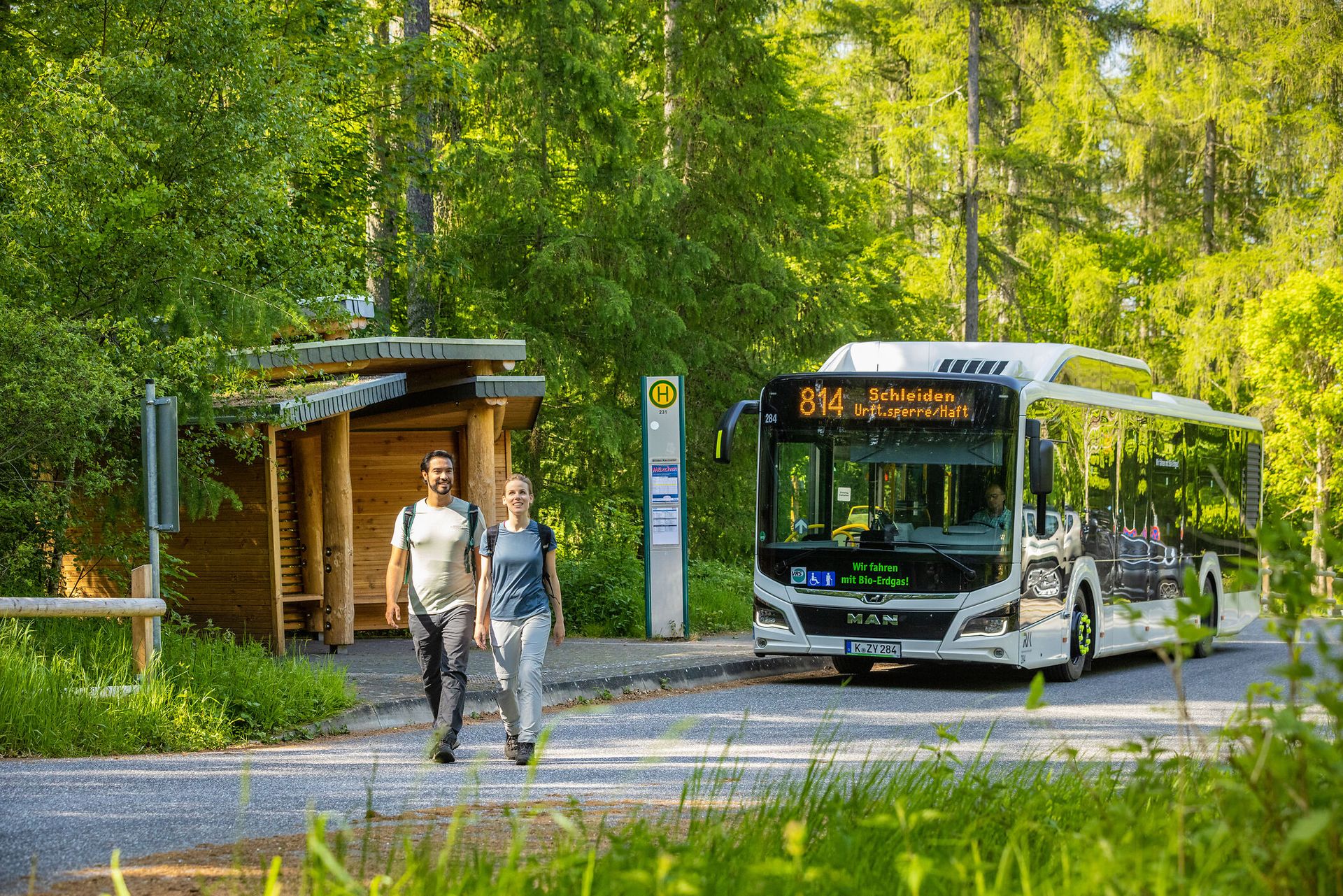 Ein moderner Linienbus hält an einer barrierefreien Haltestelle im Grünen am Wilden Kermeter; neben dem Bus ist ein hölzernes Wartehäuschen, umgeben von dichtem Wald und sommerlichem Licht.