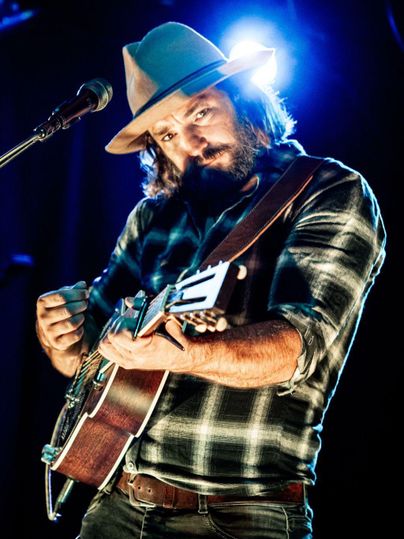 A musician performs on stage. He is wearing a hat and playing guitar while looking at the camera.