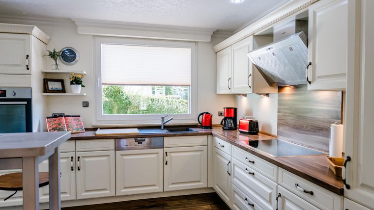 Een moderne keuken met witte kasten en een houten werkblad. Er is een gootsteen, keukenapparatuur en veel licht door het raam.