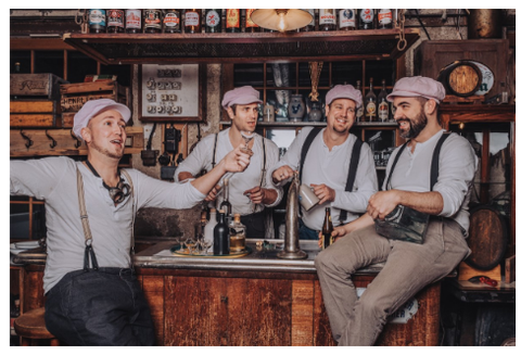 Eine Gruppe von vier Männern steht an einer Bar, alle in ähnlicher Kleidung mit Kappen und Hosenträgern. Sie scheinen Spaß zu haben und miteinander zu sprechen.
