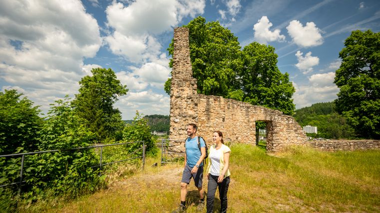 Zwei Wanderer gehen an einer alten Steinmauer vorbei, umgeben von Bäumen und blauem Himmel.
