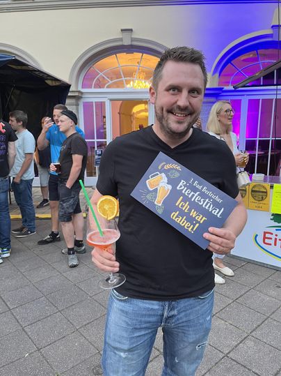 Een glimlachende man houdt een drankje vast en toont een bord met de tekst "Bierfestival - ik was erbij!". Op de achtergrond zijn andere mensen te zien op een evenement.