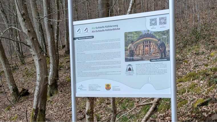 Een informatief bord over de Kolbenbergweg in een bosgebied. Het toont een afbeelding van de historische Kolbenbrug en bevat informatie over de omgeving.