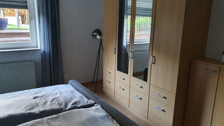 A cozy bedroom with a bed, a wardrobe, and a floor lamp. The window lets in plenty of daylight.