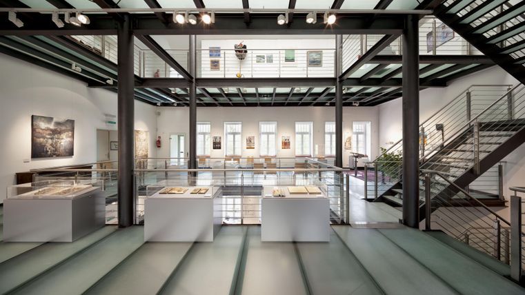 Ein modernes Kunstmuseum mit offenem Raum und großen Fenstern. Im Vordergrund stehen weiße Ausstellungsplattformen und im Hintergrund sind Bilder an den Wänden zu sehen.